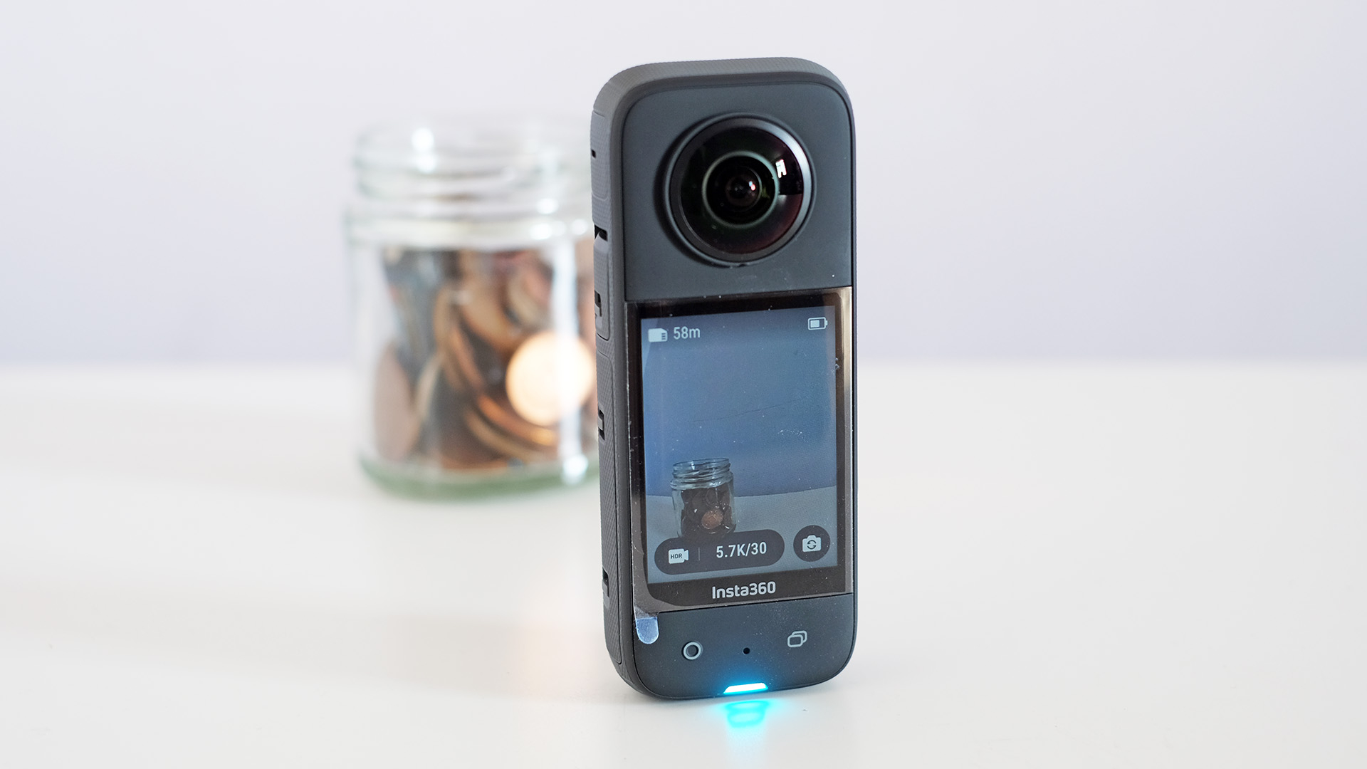 The Insta360 X3 stood on a white desk taking a photo of a jar of coins