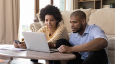 A young couple sit on the floor in front of their sofa and look at a laptop together.