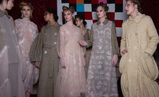 Seven female models wearing looks from Simone Rocha's collection. Five models are wearing pieces featuring sheer fabric and embellishemnts. Another model is wearing a brown plaid jacket with puffy sleeves, a belt and buttons. And the seventh model is wearing a light beige buttoned coat that puffs out in sections at the bottom. All the models are wearing earrings