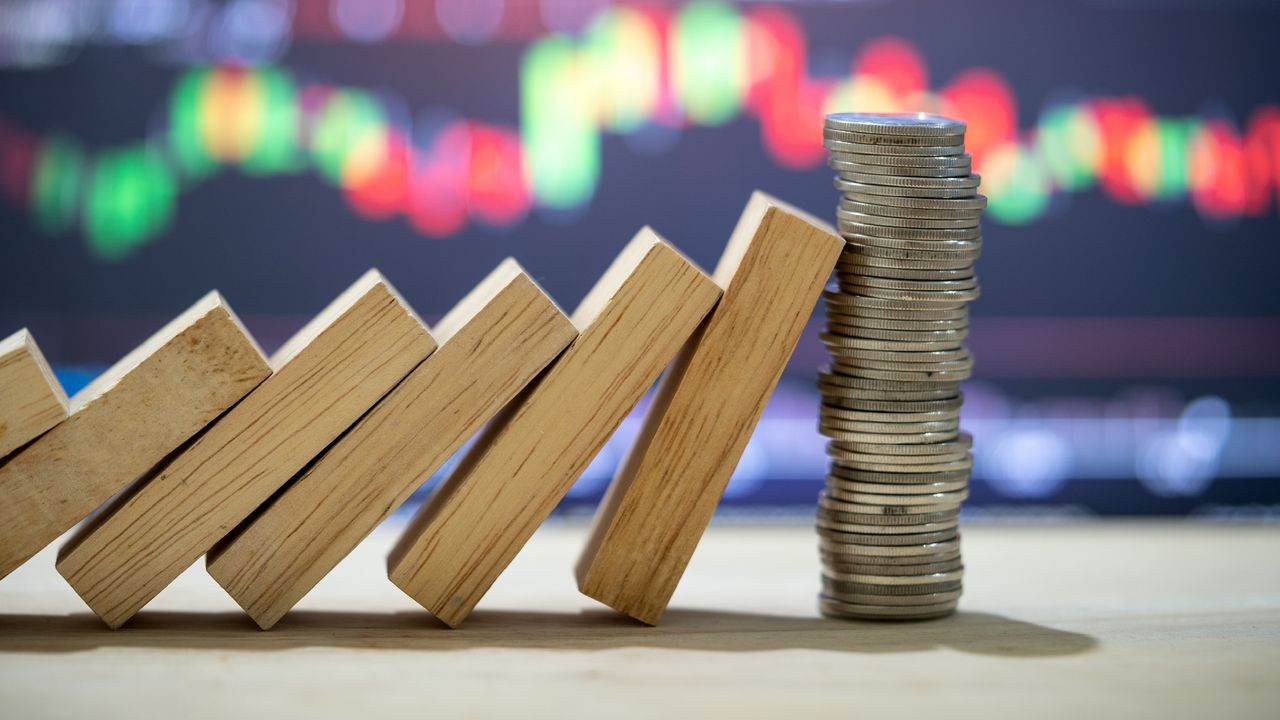 Domino-shaped blocks are tipped against a stack of coins with a trading graph in the background.