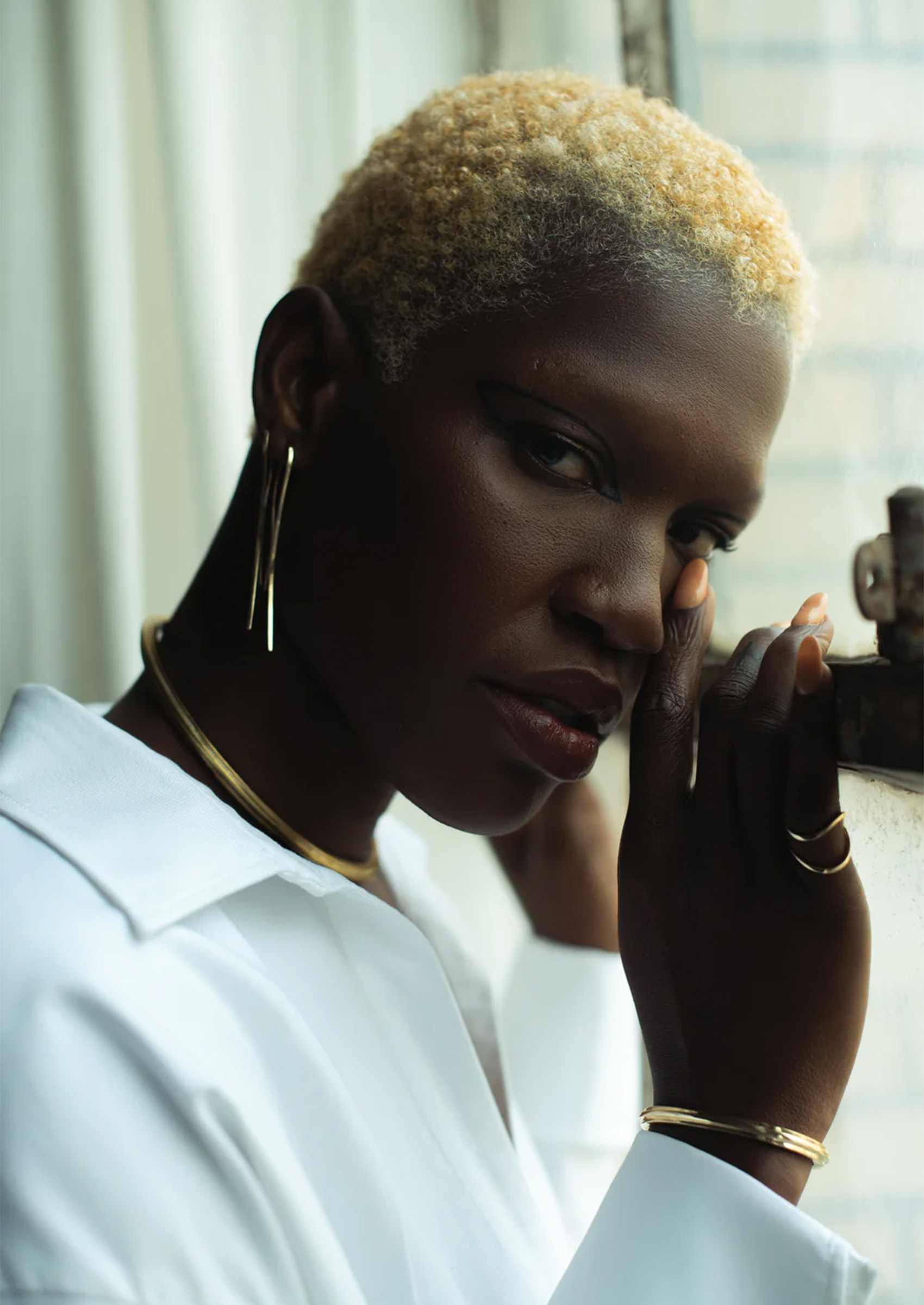 A model wearing a white shirt and V.Bellan jewelry, including gold sculptural earrings, a choker collar, bracelet, and rings.