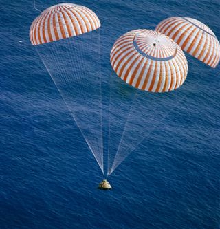Command module splashdown in the ocean. The module is supported by three large parachutes that are white and red striped.