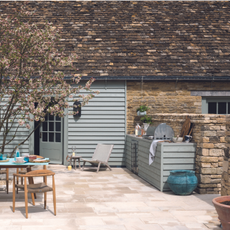 Outdoor kitchen area in garden with table and wooden chairs on patio area behind house with blue panels