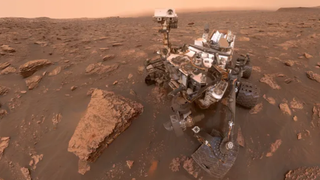 image of a large whitish rover on the surface of mars, surrounded by rocks and red dirt.