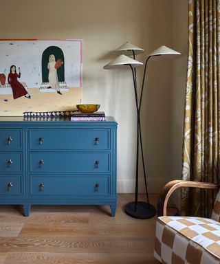 beige painted bedroom with a blue large chest of drawers and statement art, a checkerboard armchair sits in the corner