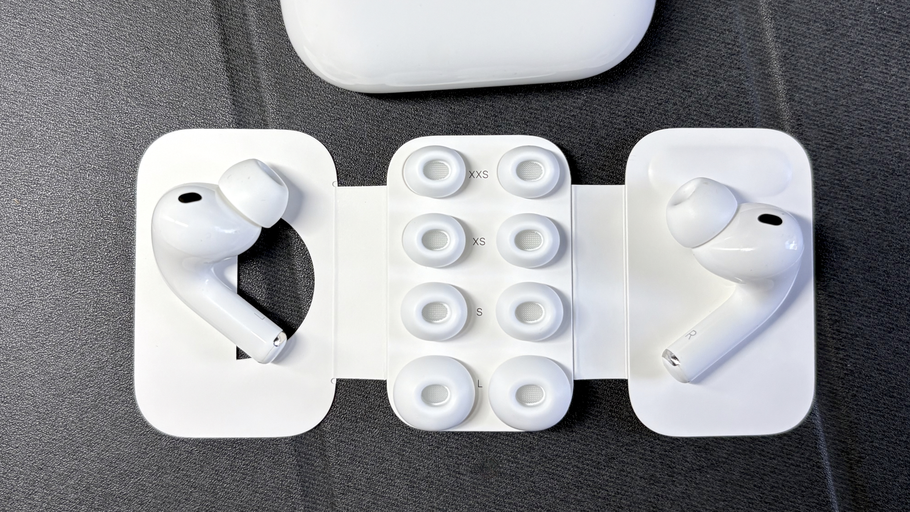 Apple AirPods Pro 3 either worn by a man standing in front of a red-brick wall, or on a table with gray twine cover