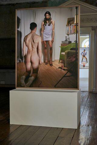 A naked male on his knees with his back to the camera, facing a female standing up wearing a short white dress. The room has a chair, books, wood floor and white curtains.