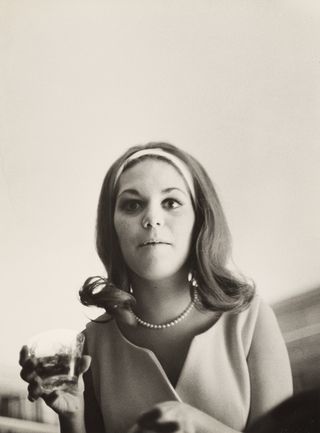 A black and white photo of a woman with shoulder length hair wearing a headband, pearls and a light coloured outfit. She is holding a glass of drink