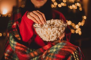 A woman eating popcorn while cosying up in a tartan blanket to watch a Christmas film