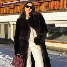 Anna LaPlaca wearing a long black fur coat, white top, white Levi's jeans, and a burgundy bag.