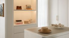 A warm white walk-in wardrobe with a display shelf with warm lighting for bags and books, a dresser island with a woven hat and a textured bowl and tall cupboards with gold hardware