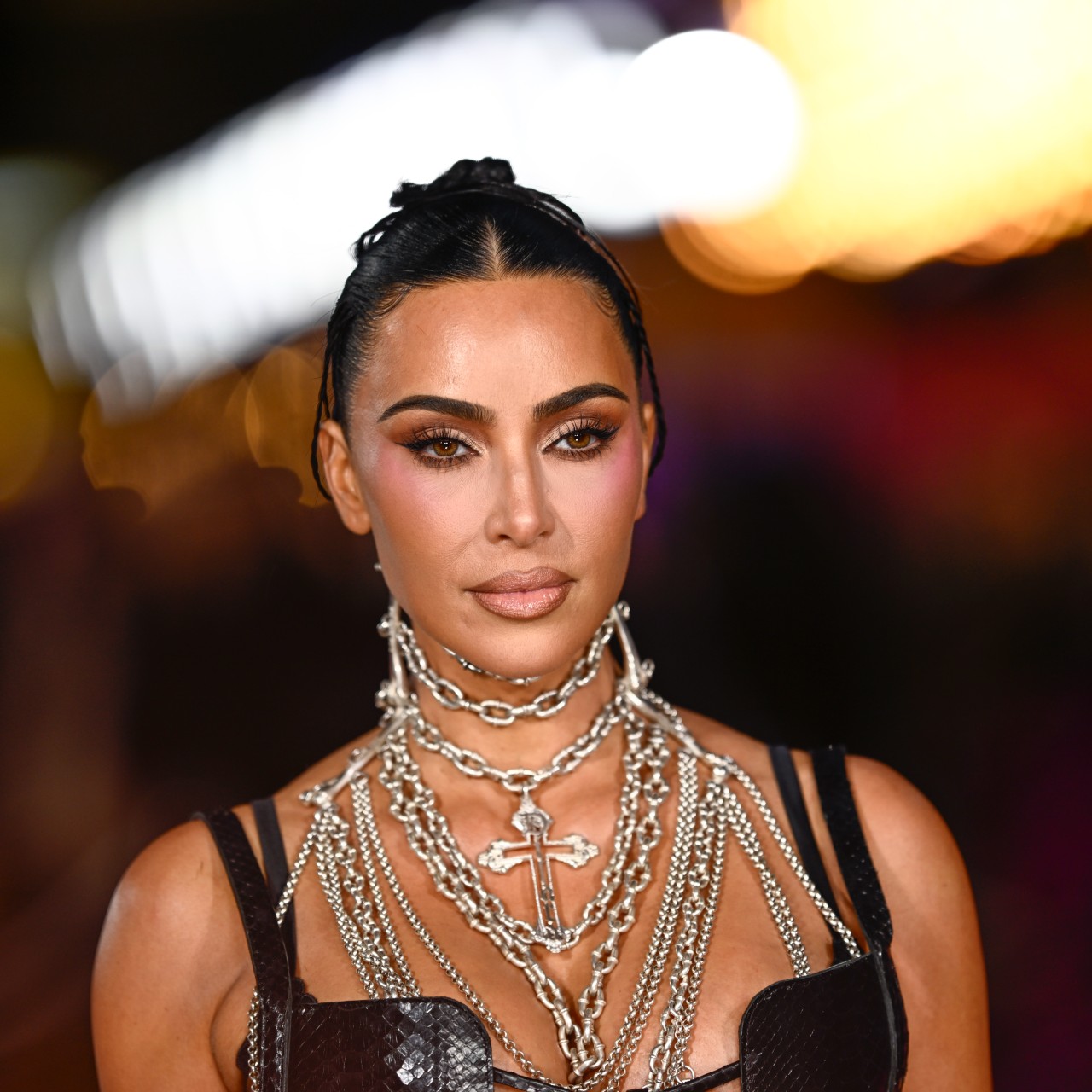 A woman with dark hair pulled back into a bun. She's wearing several silver chains around her neck and posing for a paparazzi photo on a red carpet.
