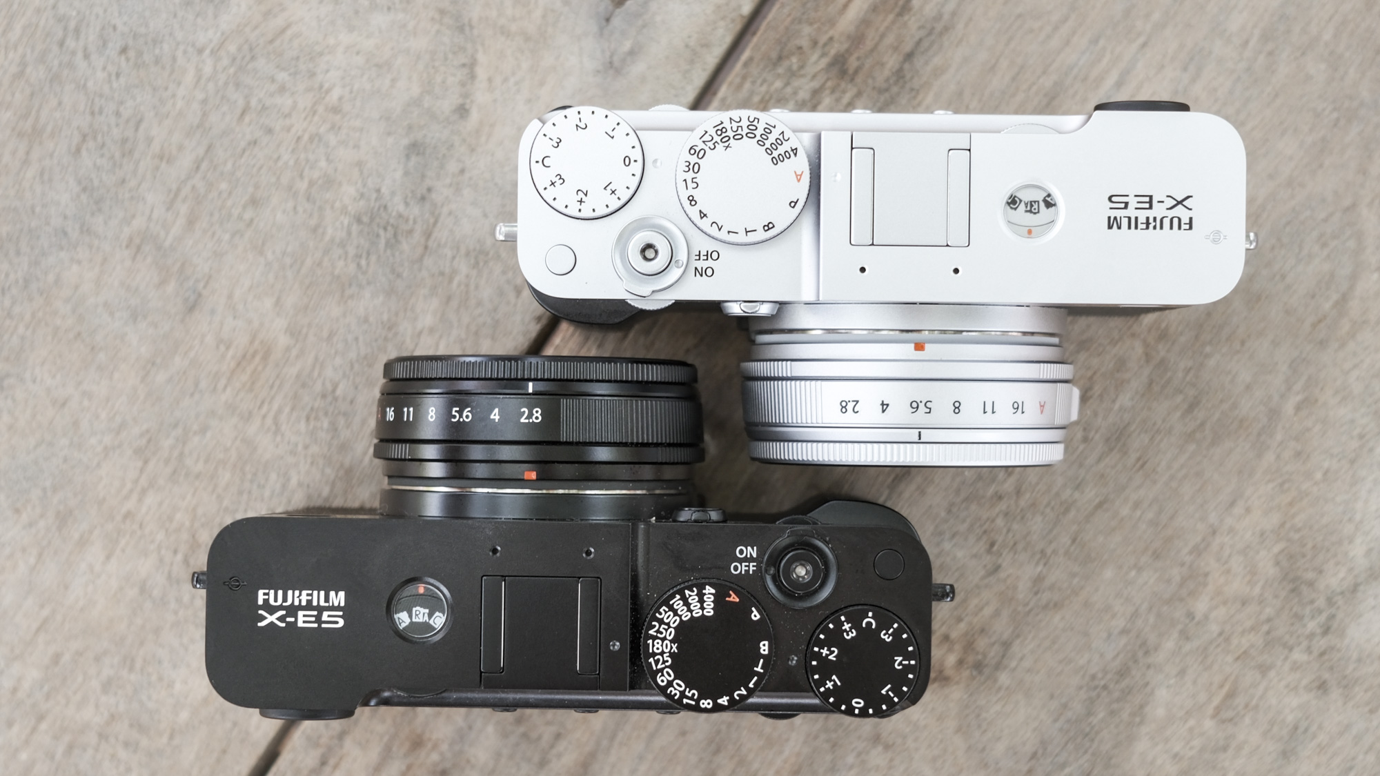 Top down of all black Fujifilm X-E5 alongside silver Fujifilm X-E5, on a wooden bench