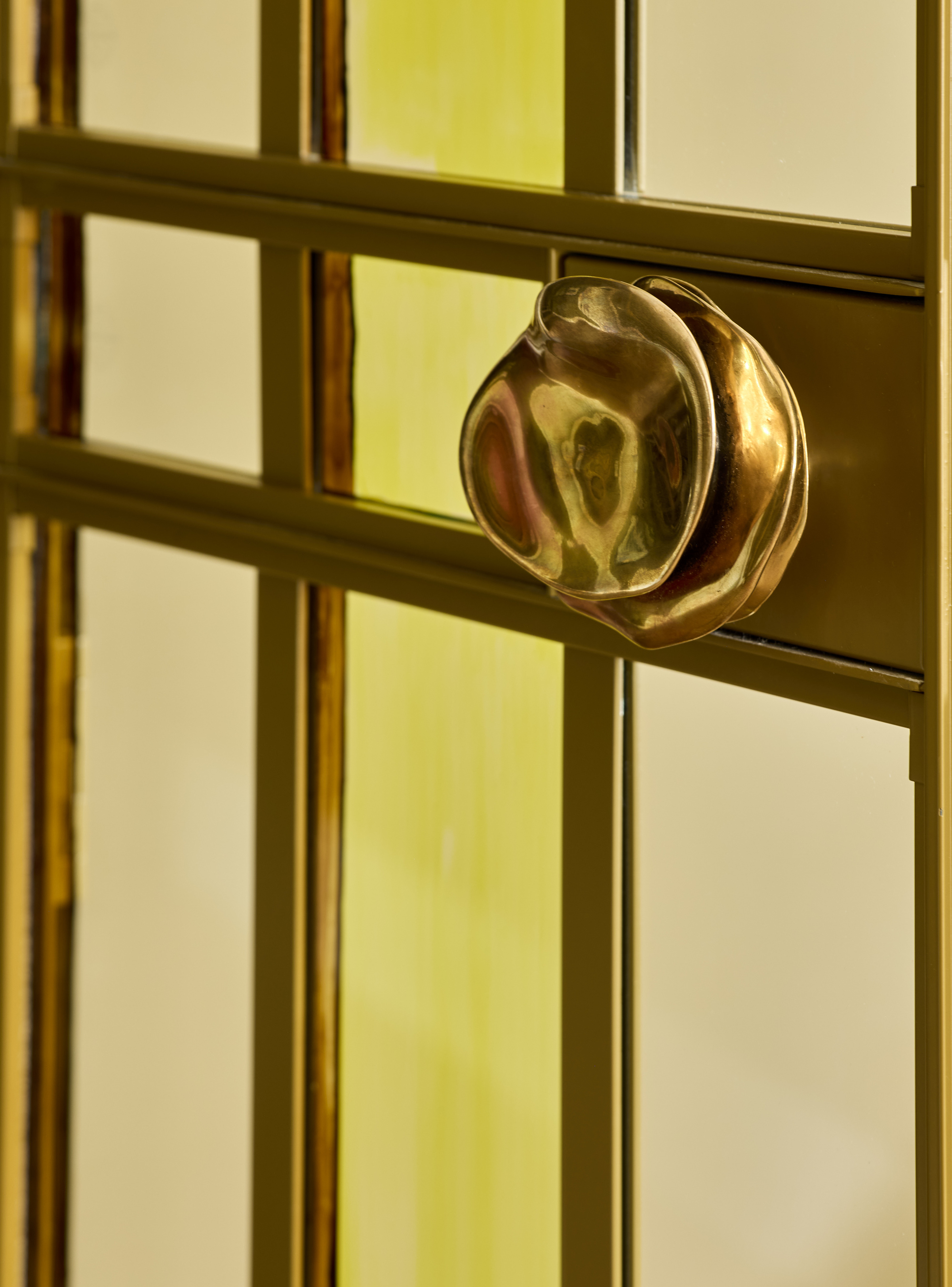 Close-up image of a gold, abstract cabinet pull in a door with gold-framed paneled glass.