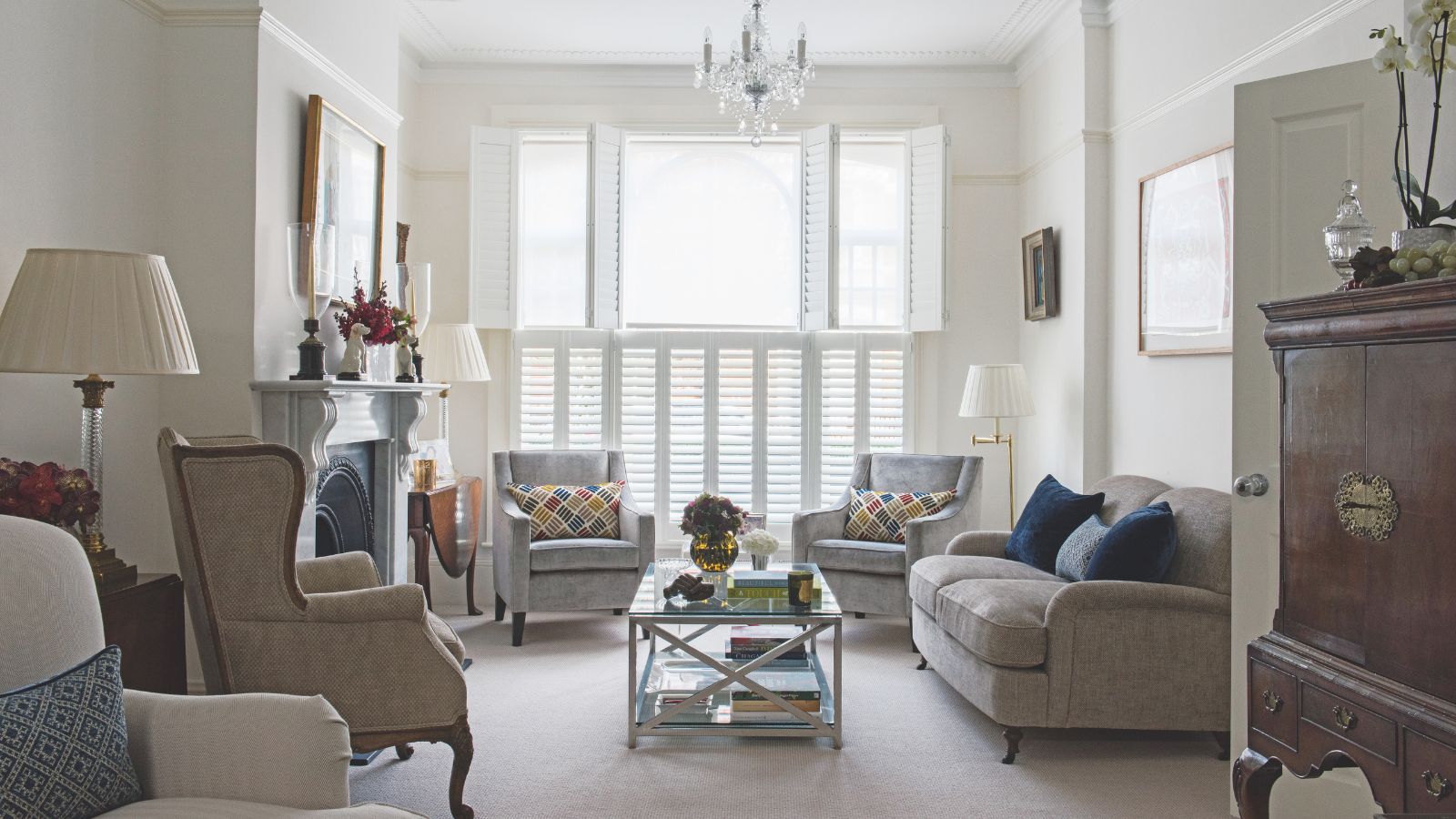 A large living room with white walls, neutral upholstered couch and chairs, a glass coffee table, large mantelpiece with artwork above, and a large wooden cabinet to the right in the foreground. In the background are large windows with white shutters.