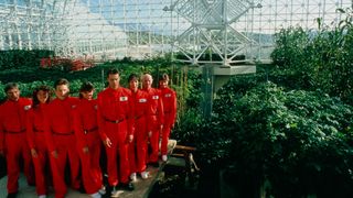 A still from Spaceship Earth in which 8 people in red boiler suits pose inside a huge greenhouse filled with plants.