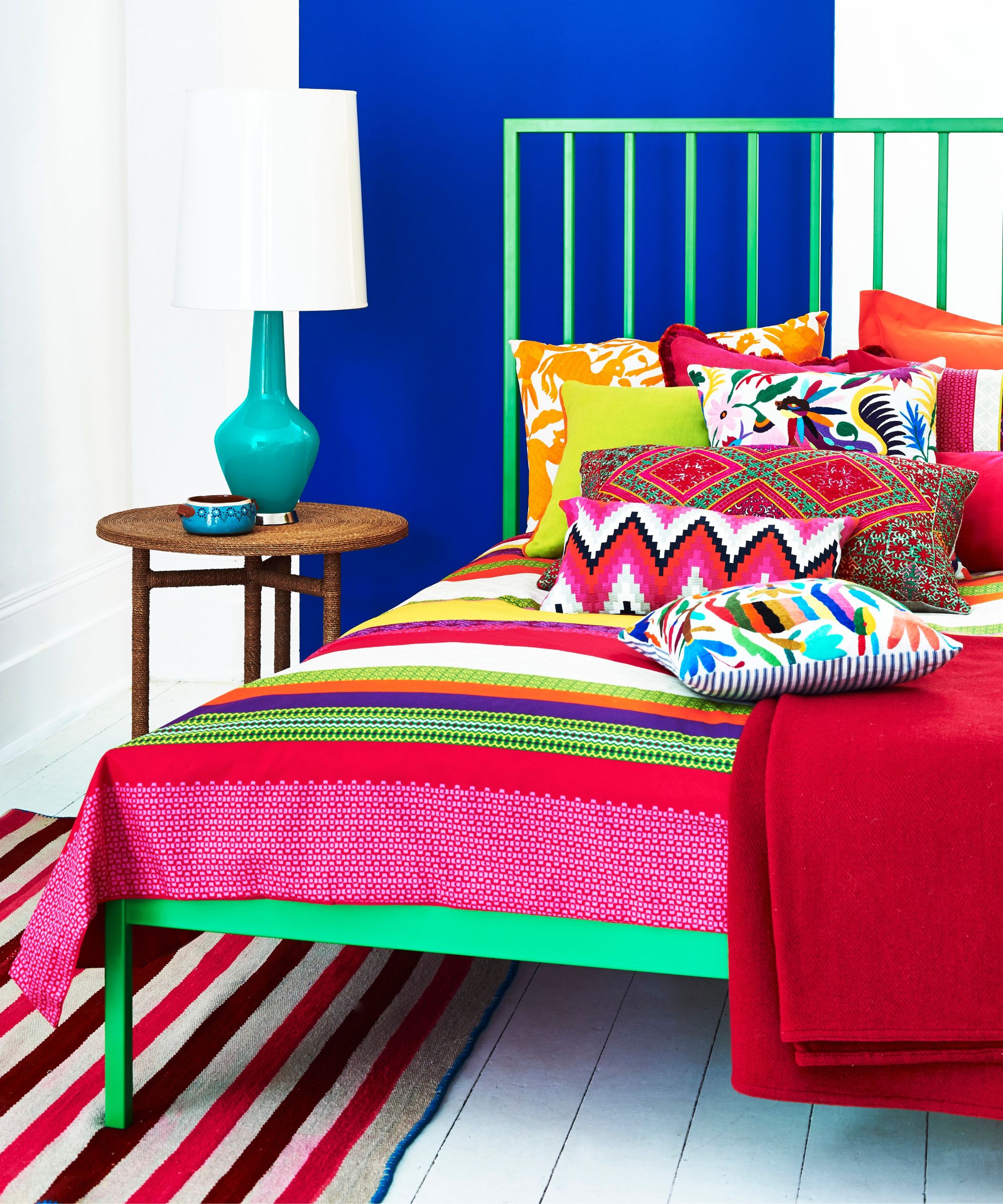 Bright bedroom with blue wall, green bed, brightly patterned bedding and cushions in assorted colors, round bedside table with teal lamp and white lampshade, and red, brown, and white striped rug.
