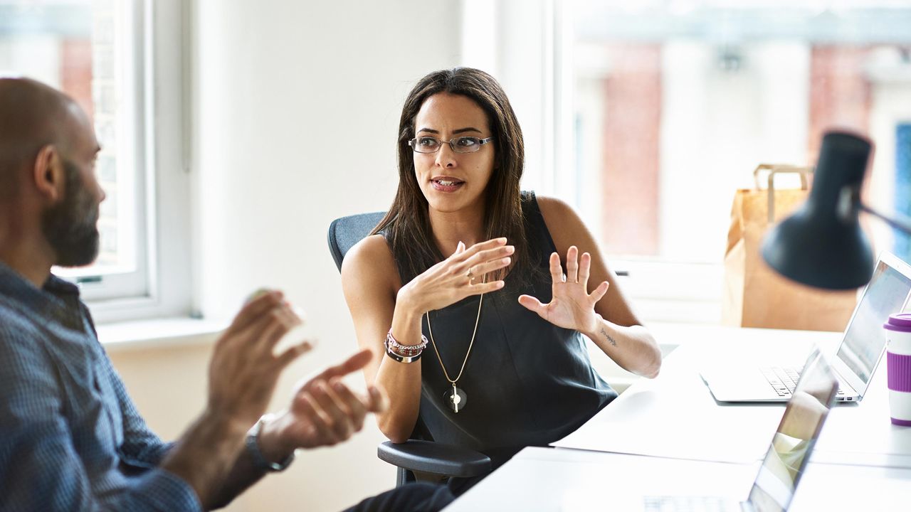 A man and woman sit at a table in a modern workspace talking together.