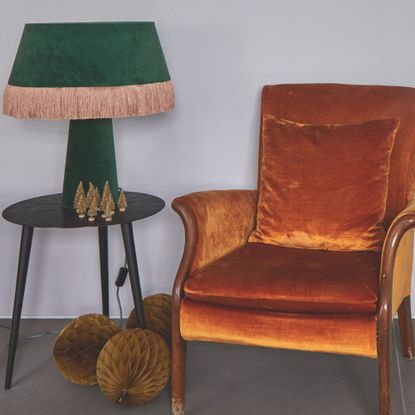 A green velvet table lamp with pink fringed trim on a small side table