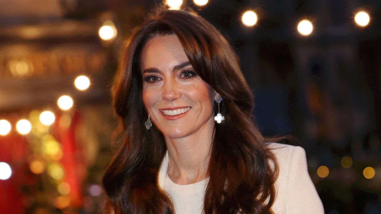 Kate Middleton wears silver earrings and a white sweater while wearing her long brown hair down and wavy