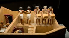 Close-up of a wooden diorama showing five human figurines. Two are facing away from the camera, and three face the camera. They are carrying sacks of grain. 