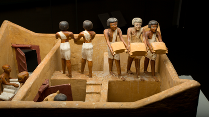 Close-up of a wooden diorama showing five human figurines. Two are facing away from the camera, and three face the camera. They are carrying sacks of grain.