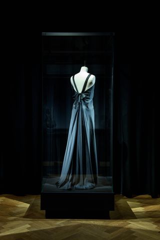 The back view of a long grey evening dress on a mannequin in a glass display case.