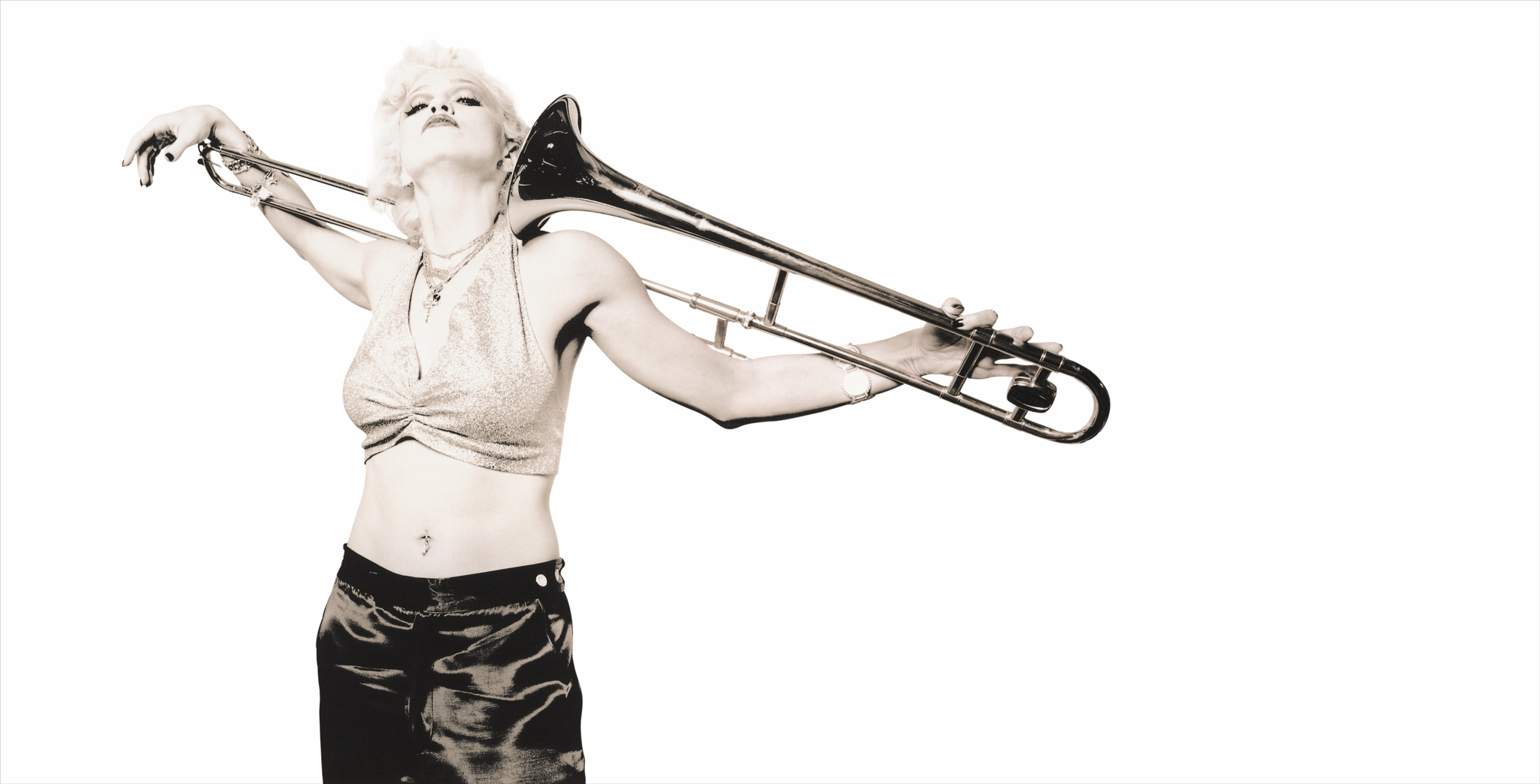 A black and white photo of Madonna, wearing a sparkling crop top and dark pants, holding a trombone across her shoulders in a dramatic pose.