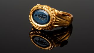 A gold ring with an oval blue gem centerpiece that has the goddess Venus carved into it. 