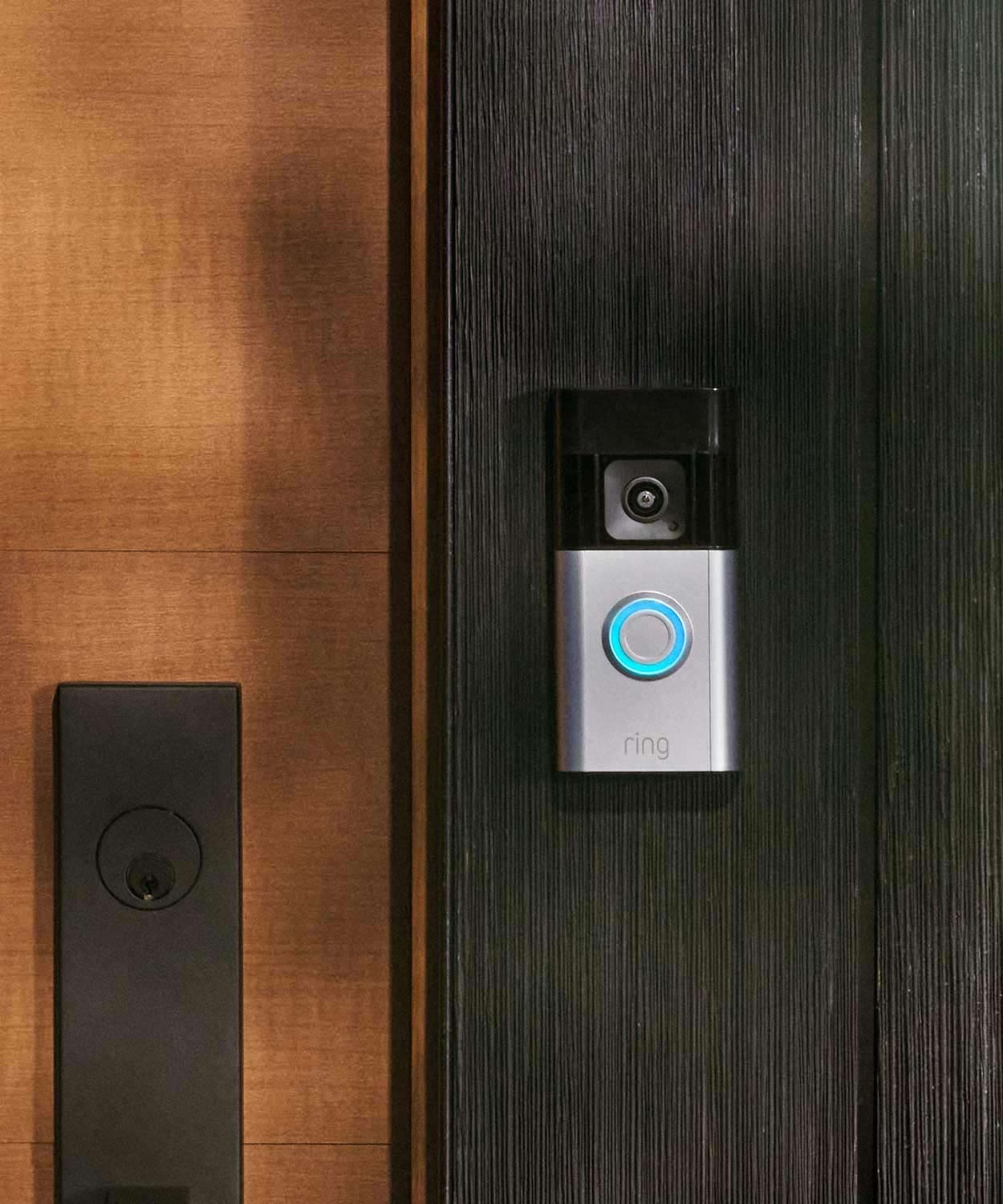 A silver and black Ring Battery Doorbell Pro mounted on a black wood wall, with a paler, warmer wood wall beside it, with a large black lock.