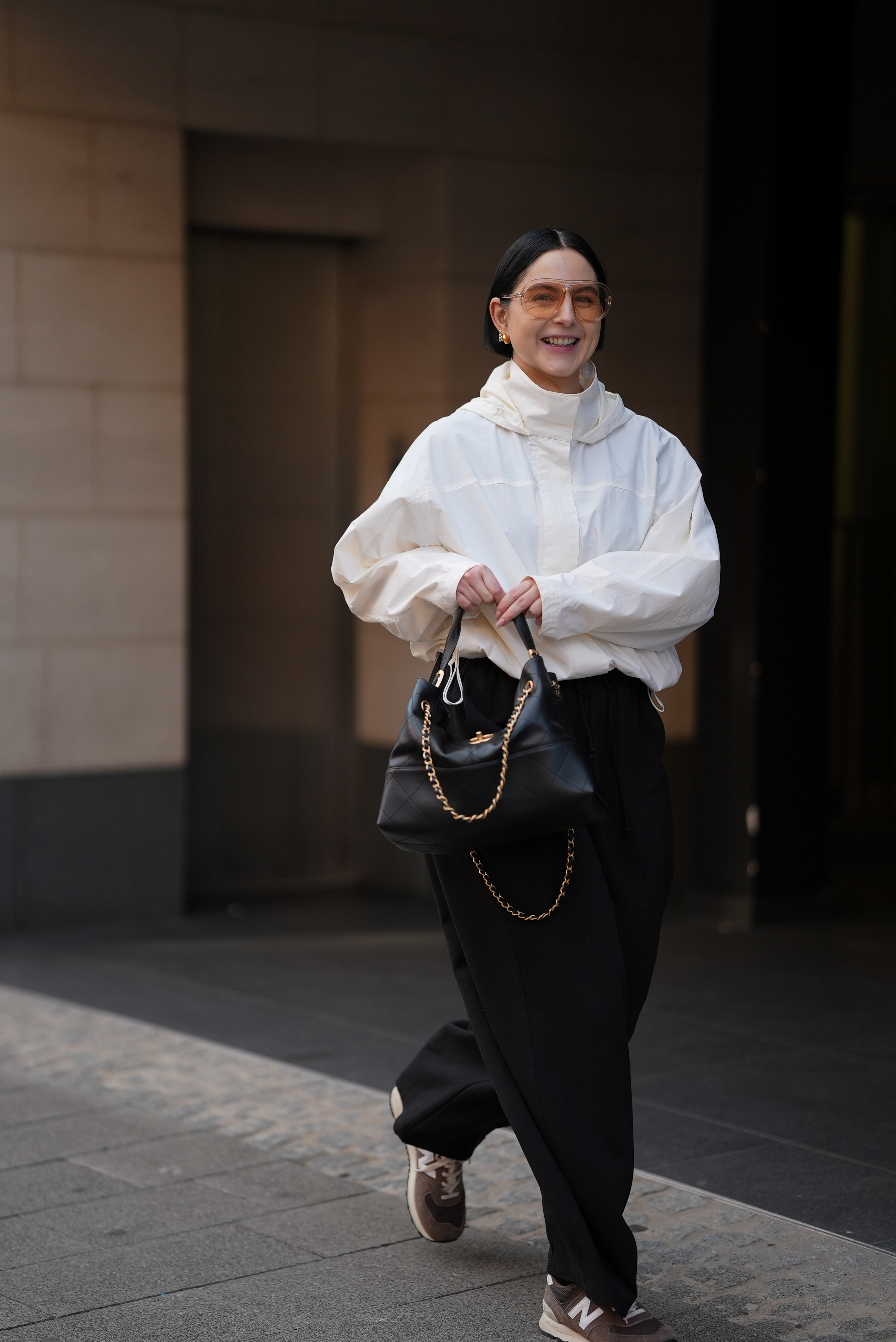 Maria Barteczko is seen wearing a white, hooded-off, oversized windbreaker-style jacket with a high collar and subtle seam detailing from UNIQLO; loose-fitting, barrel-leg black cotton trousers in a relaxed, straight leg cut from COS; athletic, brown and beige suede sneakers with white accents from New Balance; a structured, calfskin, black leather shopping bag with quilted texture and gold-tone chain details, carried by the top handle from Chanel; polished, small, gold toned hoop errings; clear, oversized aviator-style glasses with a light brown tint from TOM FORD, her shoulder-length, black hair is worn back on September 3, 2025 in Munich, Germany