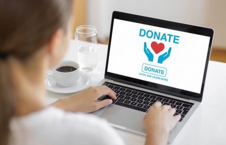 Woman using laptop with donation interface on screen showing heart and hands icons for charity support.
