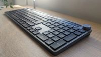A sleek, dark grey wireless keyboard with a numeric keypad and a prominent circular volume control knob, resting on a light-colored wooden desk surface.