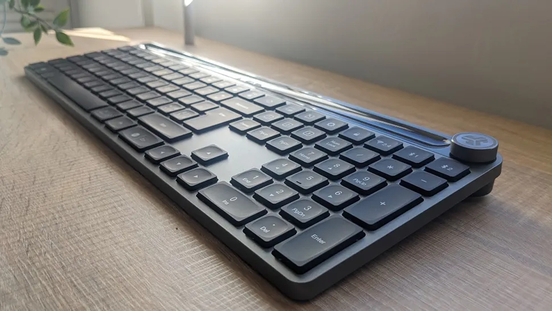 A sleek, dark grey wireless keyboard with a numeric keypad and a prominent circular volume control knob, resting on a light-colored wooden desk surface.