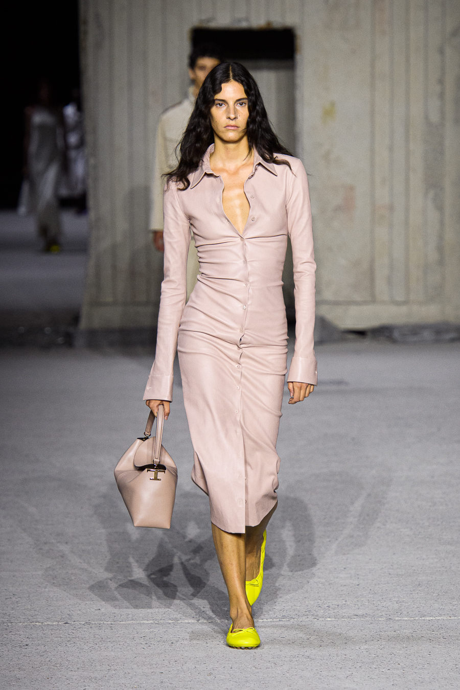 A female model wearing a light brown long sleeved long dress and yellow shoes walking down a runway.