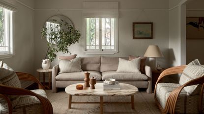 A warm living room with a pair of wood-armed sofa seaters, a beige couch, a potted tree, a marble coffee table, hanging art, and a round mirror