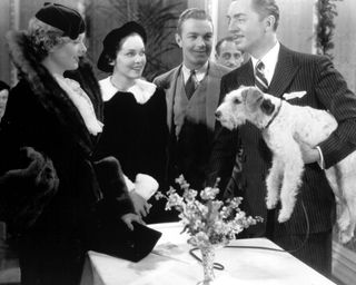 a group of people including a man holding a dog smile and stand around a table in the movie The thin man