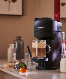 Nespresso coffee maker on a white work surface, surrounded by mugs of coffee, against beige walls.