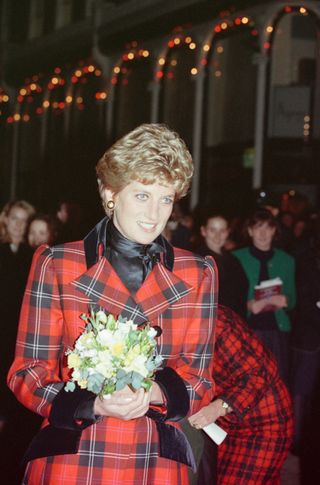 Princess Diana wearing a red tartan coat and holding a bouquet of flowers standing in front of a crowd of people and christmas lights