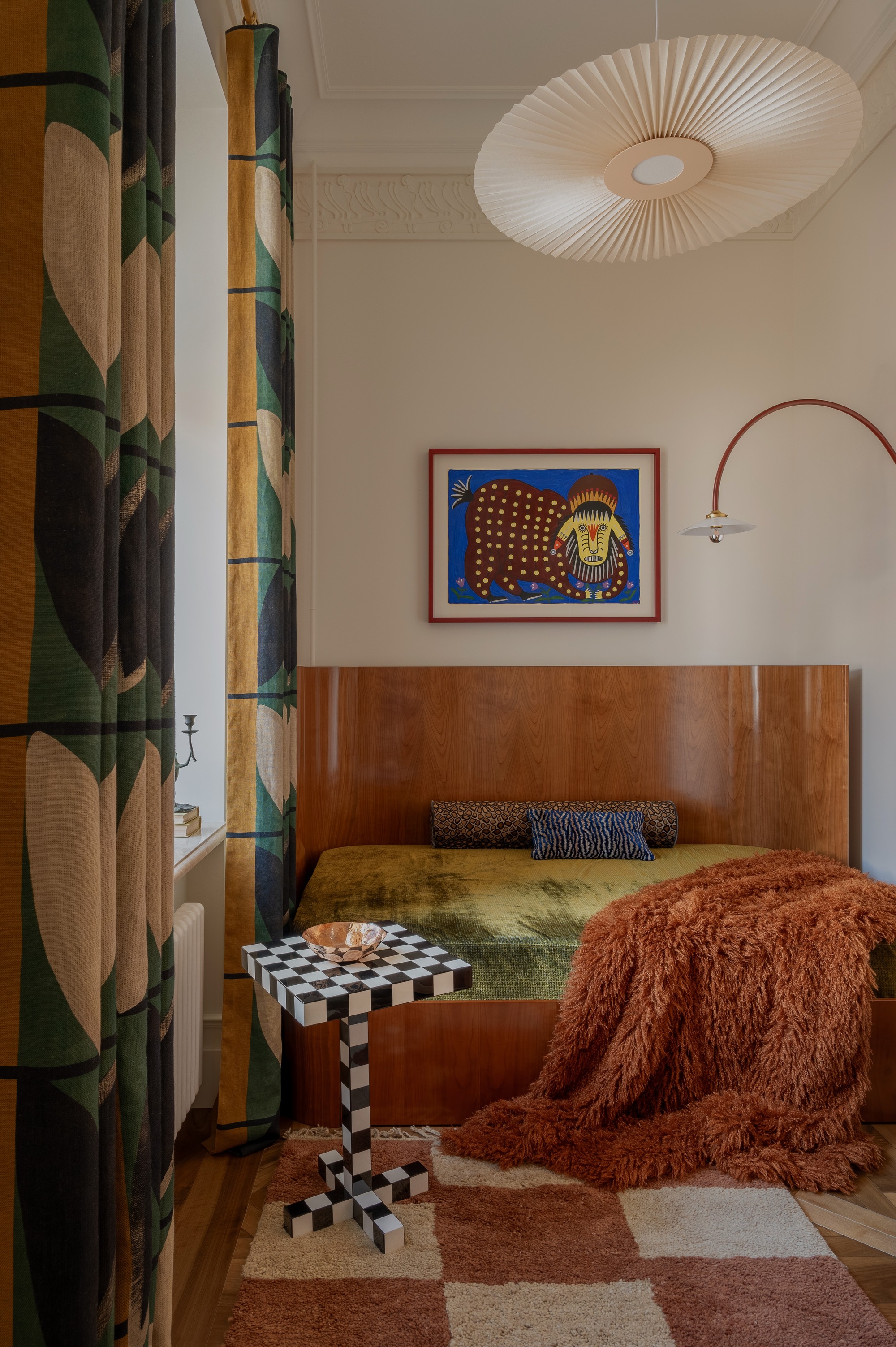 Image of a small sitting area with a velvet, olive green bench seat that has a terracotta fuzzy throw blanket draped on it. There is a black and white checkerboard side table and a terracotta and white checkerboard rug.