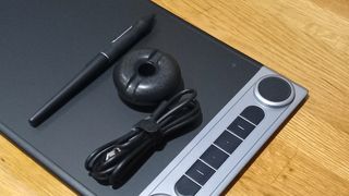 Huion Inspiroy Dial 2 review; a drawing tablet on a wooden table with a style and cable