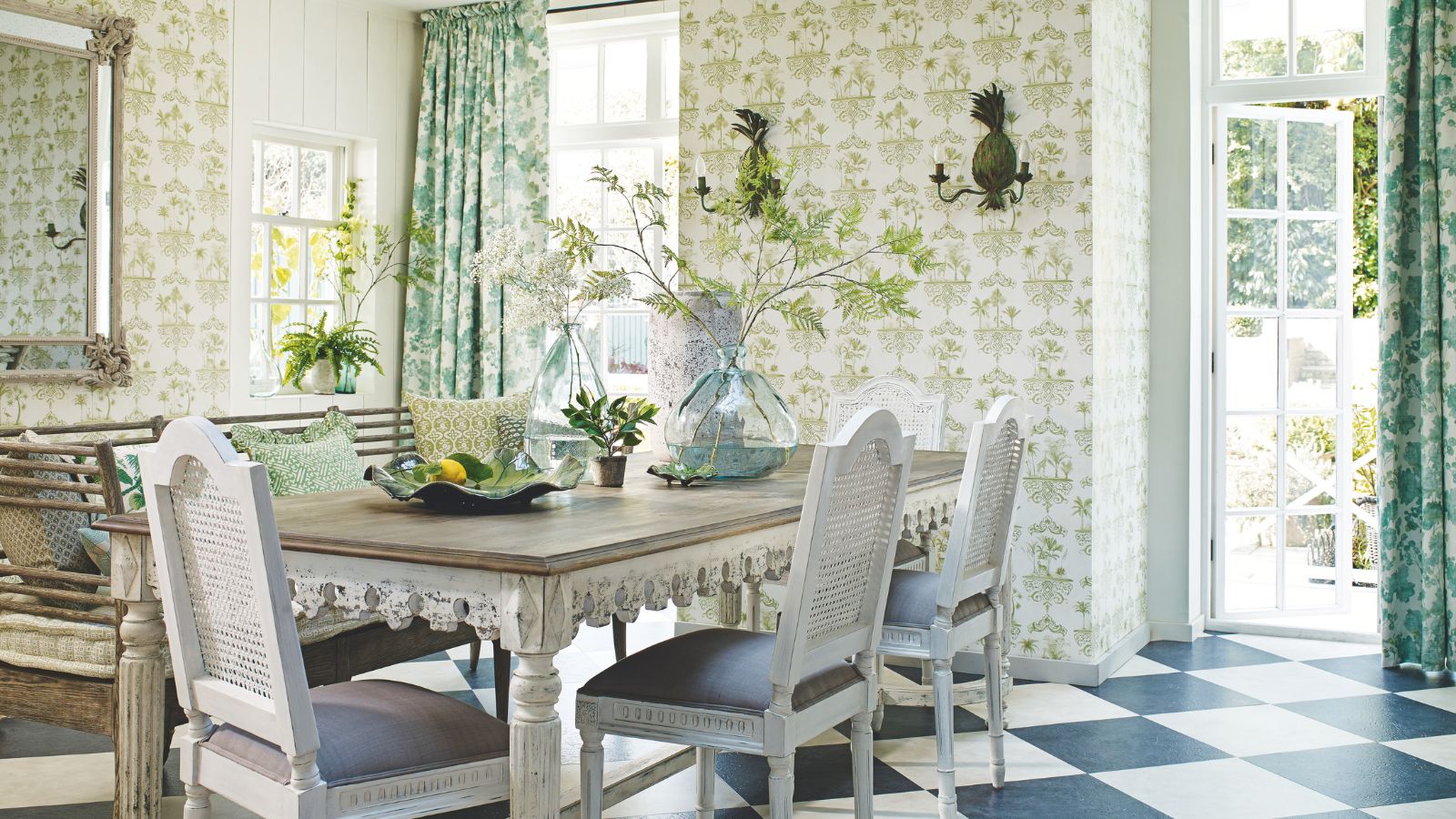 A statement dining area with green patterned wallpaper, a large dining table with white rattan chairs, green patterned curtains, black and white tiled flooring and an open door to a leafy, sunny garden. A statement mirror is seen to the right above a wooden bench with green patterned cushions.
