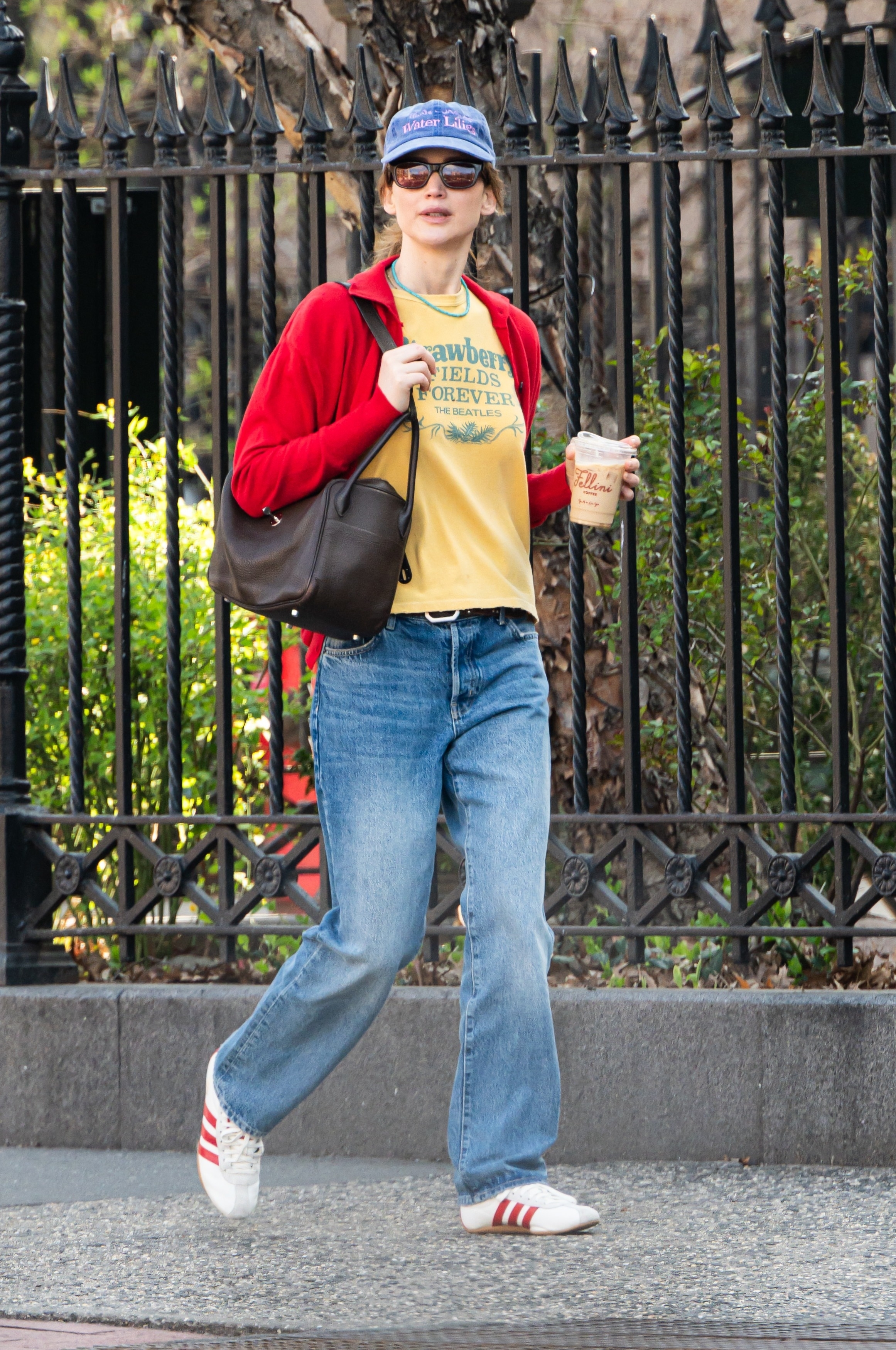 Jennifer Lawrence wearing Adidas white and red Tykyo sneakers, blue jeans, a yellow tee shirt, red sweater, blue hat, and brown bag.