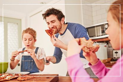 father and children stand in kitchen eating slices of pizza