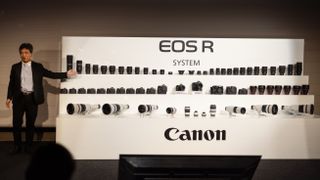 Canon executive Manabu Kato stands in front of a display showcasing the entire EOS R range of cameras and RF lenses