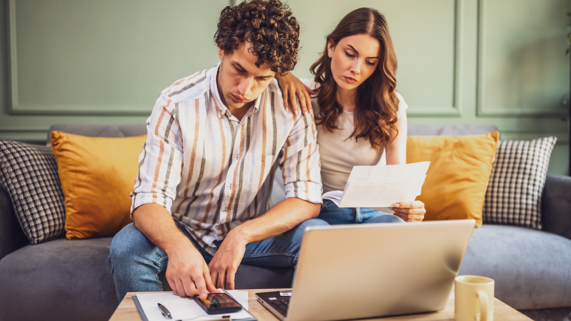 Couple sitting on their living room couch together and reviewing financial documents using a calculator and laptop