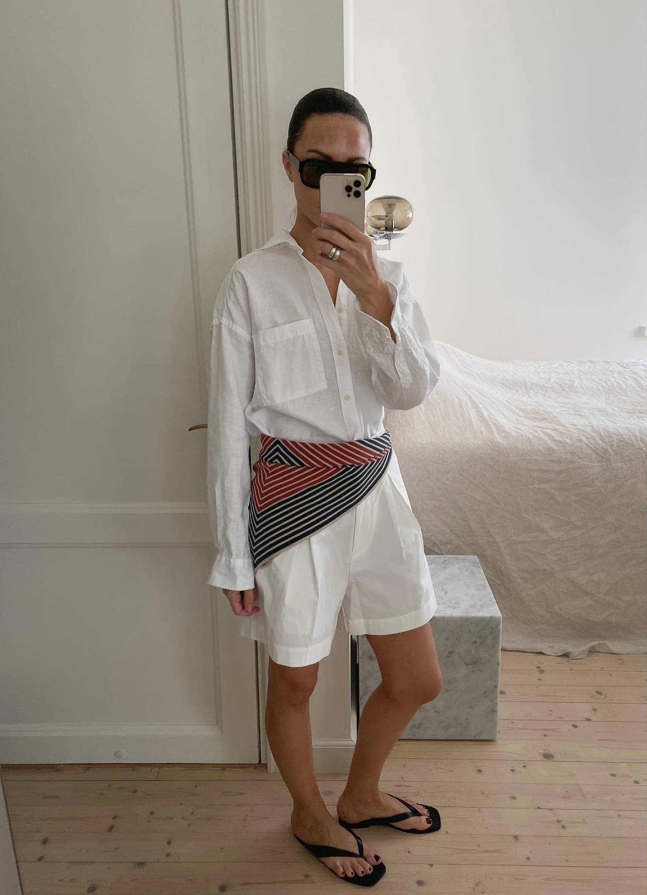 Finnish style influencer Johanna Piispa poses for a mirror selfie wearing a white button-down shirt, striped hip scarf, white shorts, and black flat flip-flop sandals