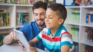 Smiling male teacher reads tablet computer with smiling 7-year-old boy. 