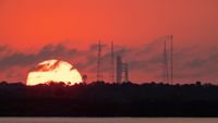 a massive-looking sun rises in a red sky behind the silhouettes of several skeletal metal towers and a rocket upright on a launch pad