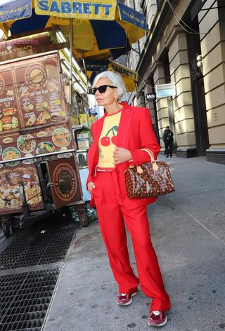 A woman wearing a bright red blazer with matching trousers styled with a yellow cherry-print sweater, a Louis Vuitton bag, and red chunky sneakers.